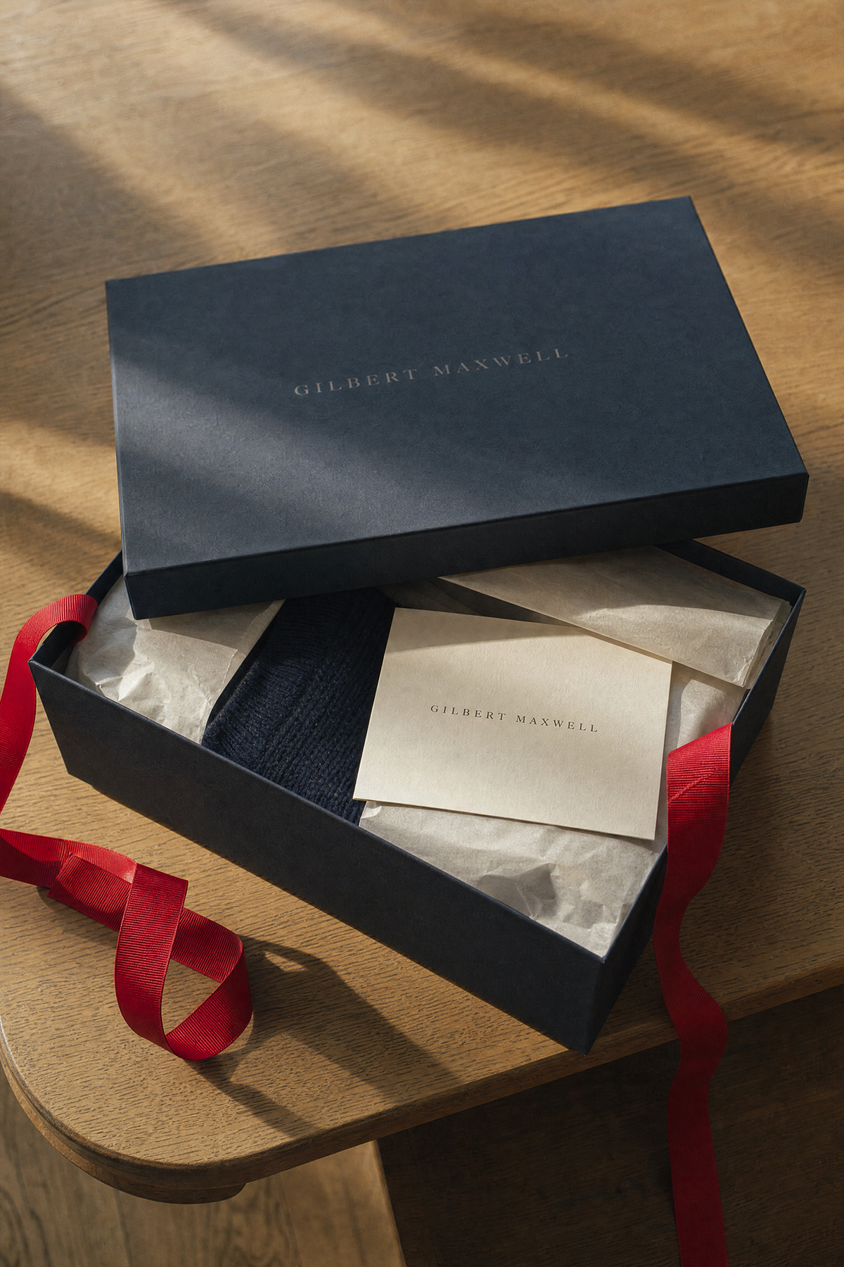A navy knit folded in tissue paper inside an open Gilbert Maxwell box, the lid resting on top and a red ribbon beside it